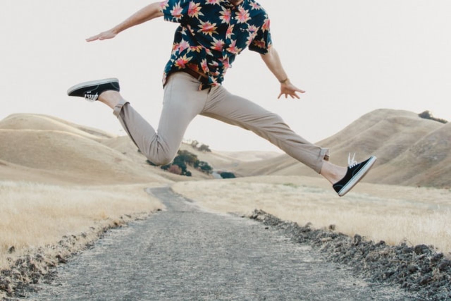 A person in a floral shirt leaping along a gravel path through dry, rolling hills