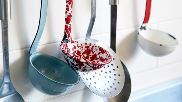 Enamel ladles and a slotted spoon hanging in a row against a white tiled kitchen wall