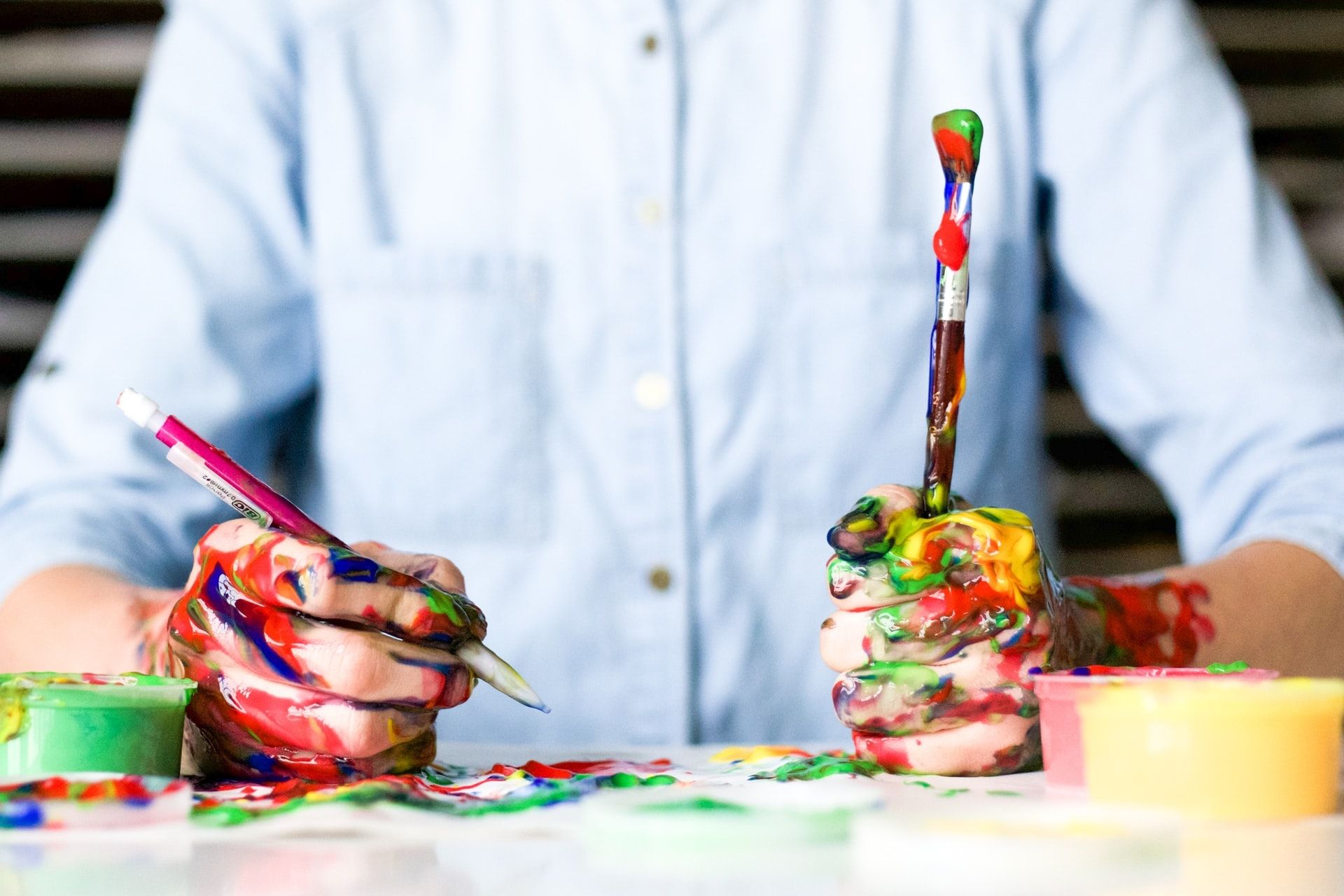 A child with paint-covered hands holding two paintbrushes over a table of paint pots
