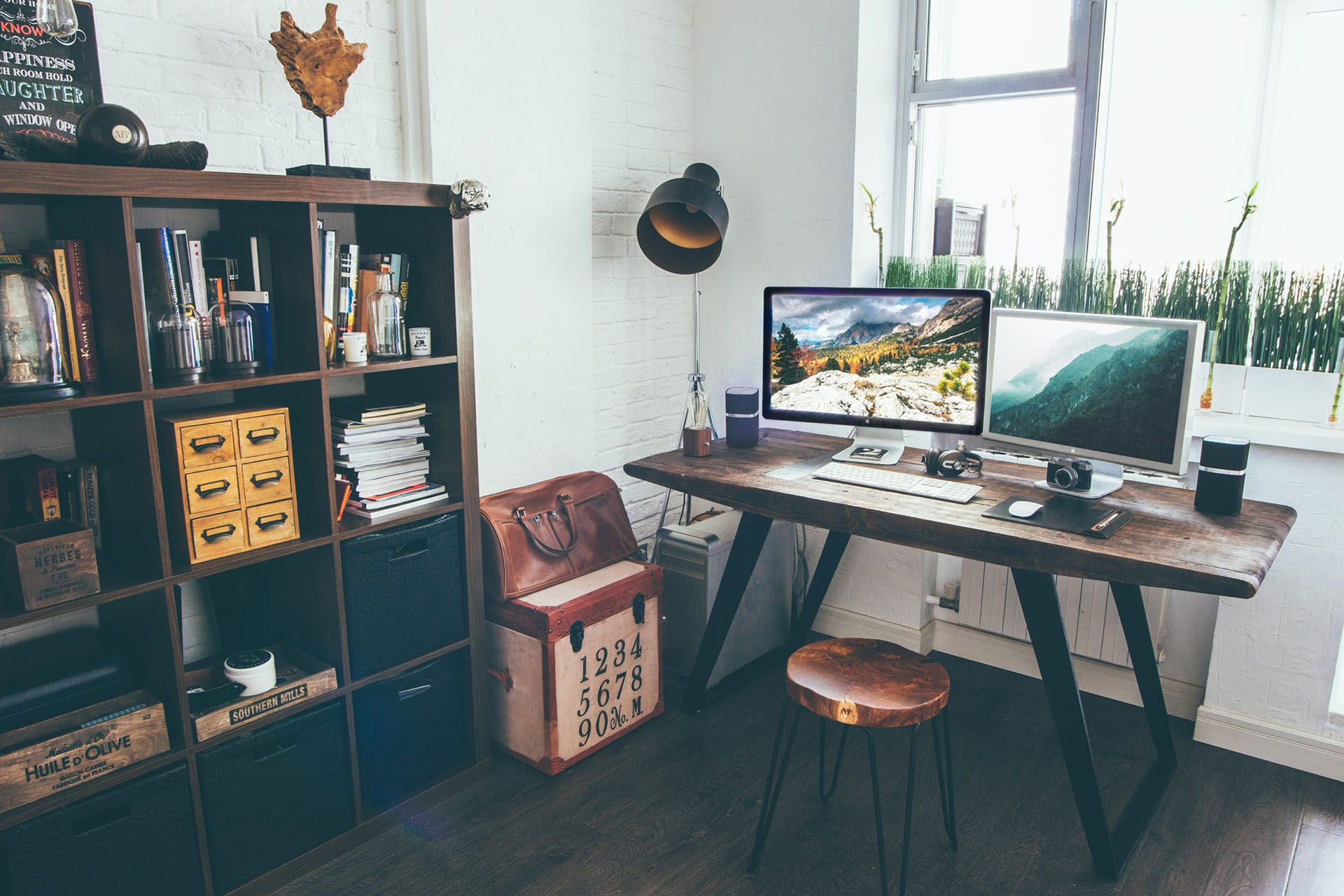 A home office with a wooden desk, two monitors, a leather bag and shelves of books and drawers