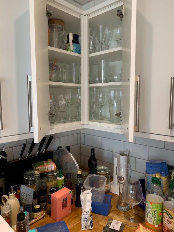 Mid-organisation of a kitchen cupboard, there are plates, bowls and other items on the kitchen counter below, as well as a large quantity of bottles on a cupboard shelf, an empty cupboard shelf, and a pile of miscellaneous containers on another cupboard shelf