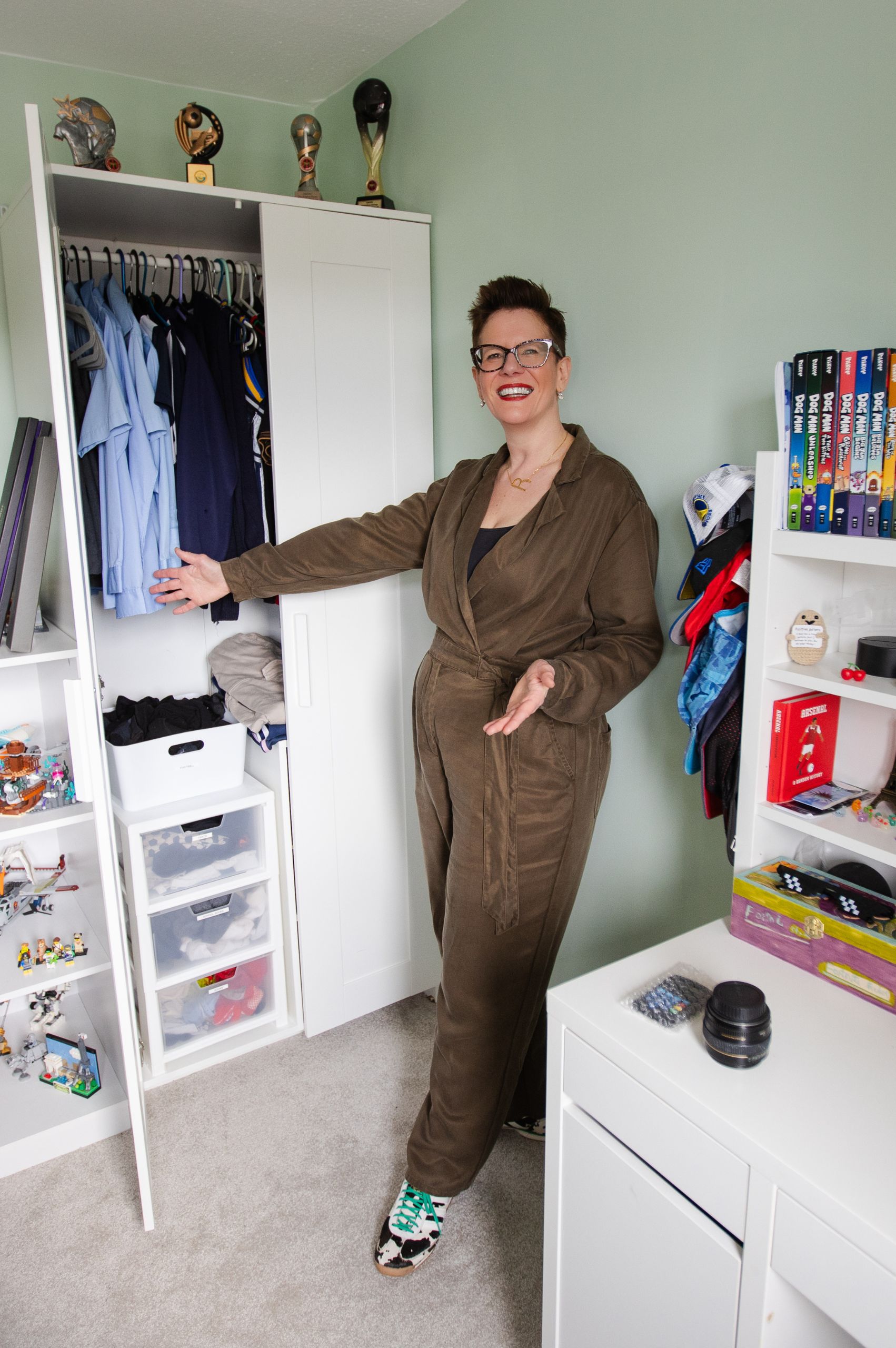 Roxy of Declutter & Reorganise with Roxify smiling and gesturing proudly toward a neatly organized open white wardrobe in a child's bedroom. She is wearing a brown jumpsuit and glasses, standing on a grey carpet surrounded by tidy white shelving units filled with arranged clothes, books, caps, and toys.