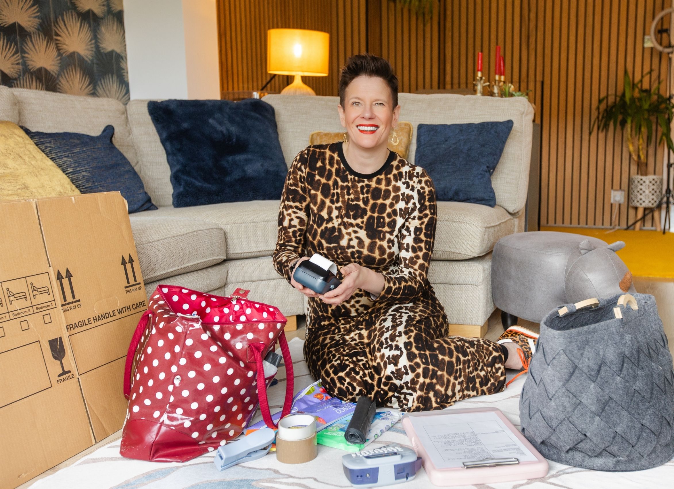 Roxy of Declutter & Reorganise with Roxify smiling and holding a label maker while sitting on a living room rug. She is wearing a leopard print outfit and is surrounded by a cardboard moving box, a red polka-dot tote bag, a grey woven basket, and various organising supplies, with a cozy sofa behind her.