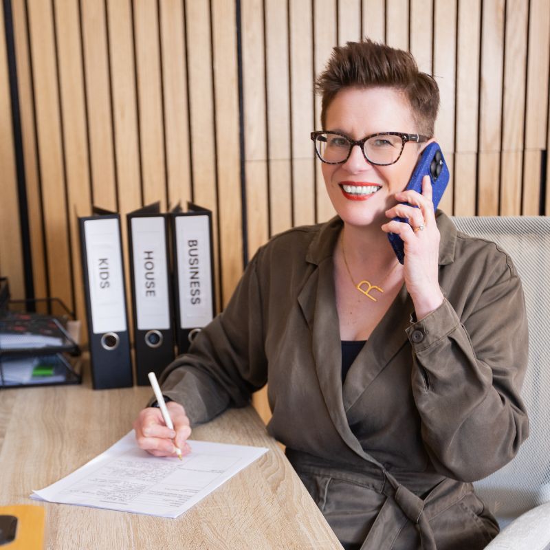 Roxy of Declutter & Reorganise with Roxify smiling and talking on a blue smartphone while sitting at her desk. She is wearing an olive green jumpsuit and glasses, holding a pen over a document. On the desk next to her are three neat black binders labeled "KIDS," "HOUSE," and "BUSINESS," set against a warm wood-slatted wall.