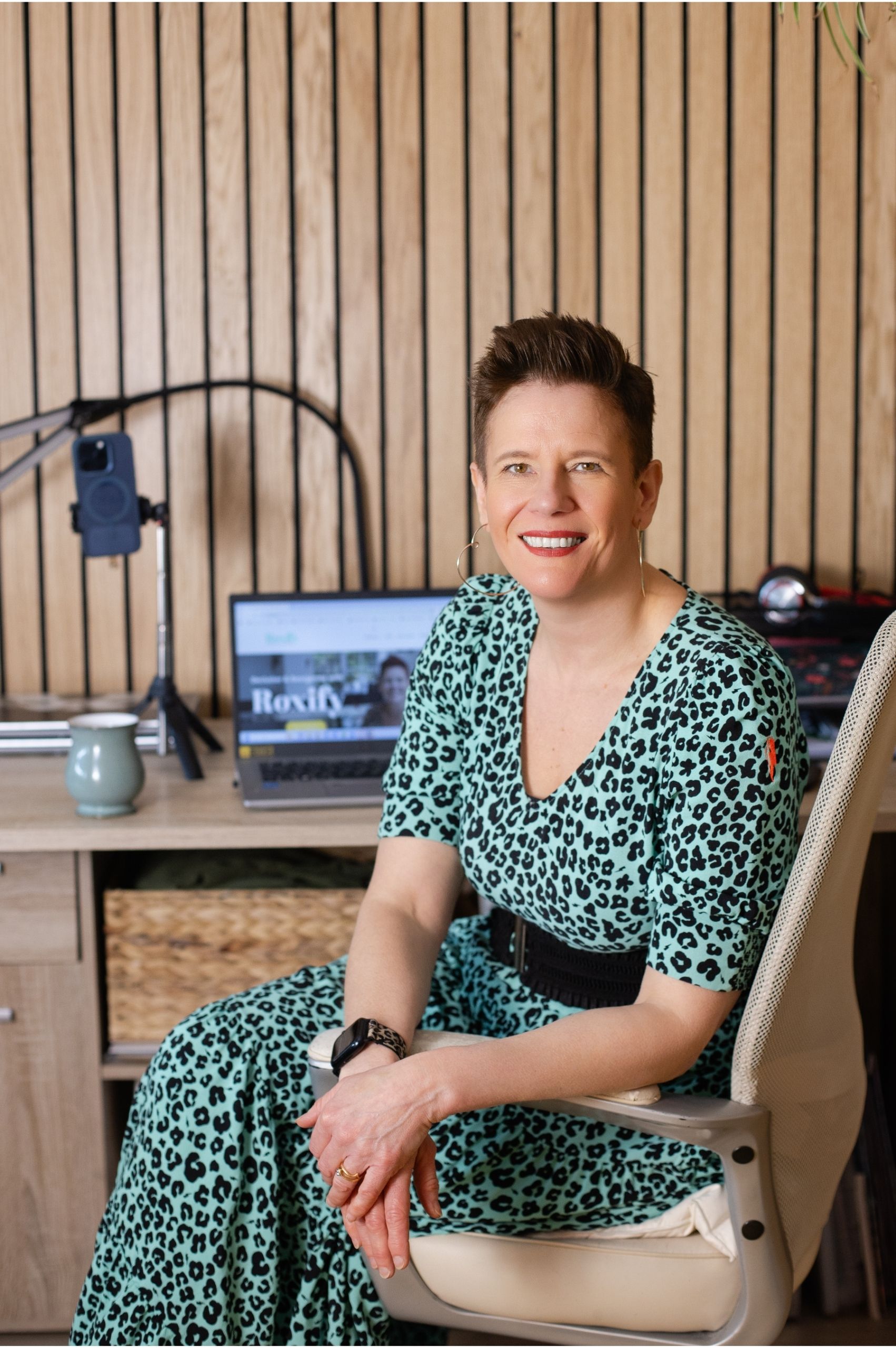 Roxy sat at her desk Roxy of Declutter & Reorganise with Roxify smiling as she sits in a cream office chair at her workspace. She is wearing a turquoise leopard print dress with a black belt. Behind her is a wood-slatted wall and a desk holding a laptop displaying her "Roxify" website, a mug, and a smartphone mounted on a tripod