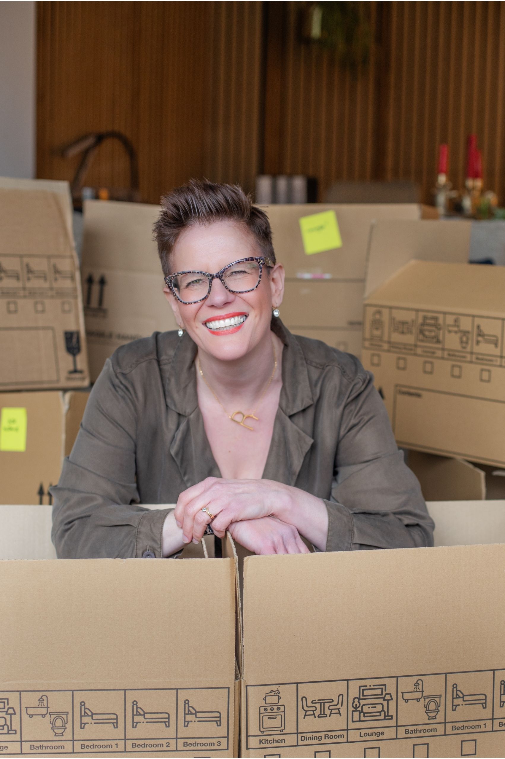 Roxy of Declutter & Reorganise with Roxify smiling brightly and leaning over cardboard packing boxes used for organising sheds and garages. She is wearing an olive green outfit and glasses, surrounded by several boxes with printed labels and yellow sticky notes.