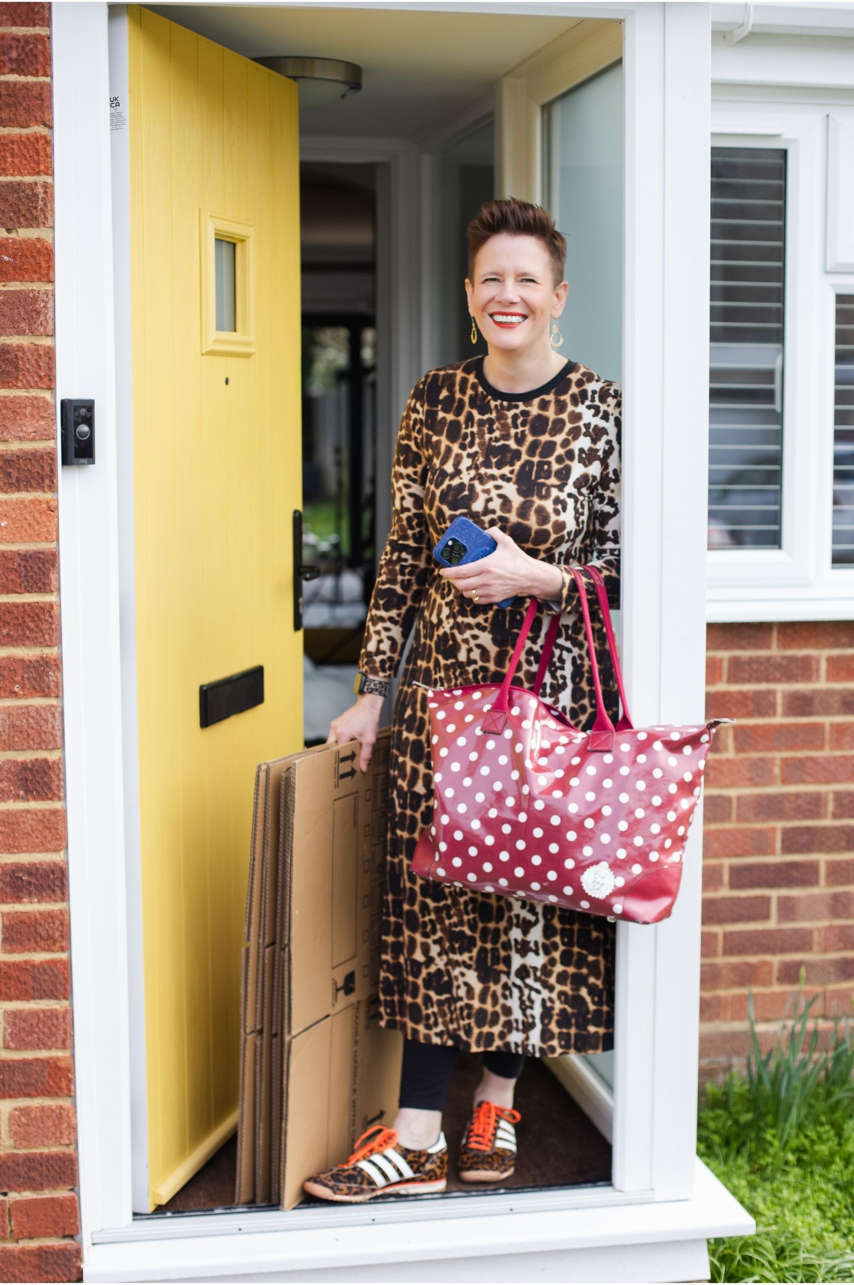 Roxy of Declutter & Reorganise with Roxify smiling as she stands in the open yellow front door of a brick house. She is wearing a leopard print outfit and trainers, and is carrying a large red polka-dot tote bag, several flat-packed cardboard boxes, and a smartphone.