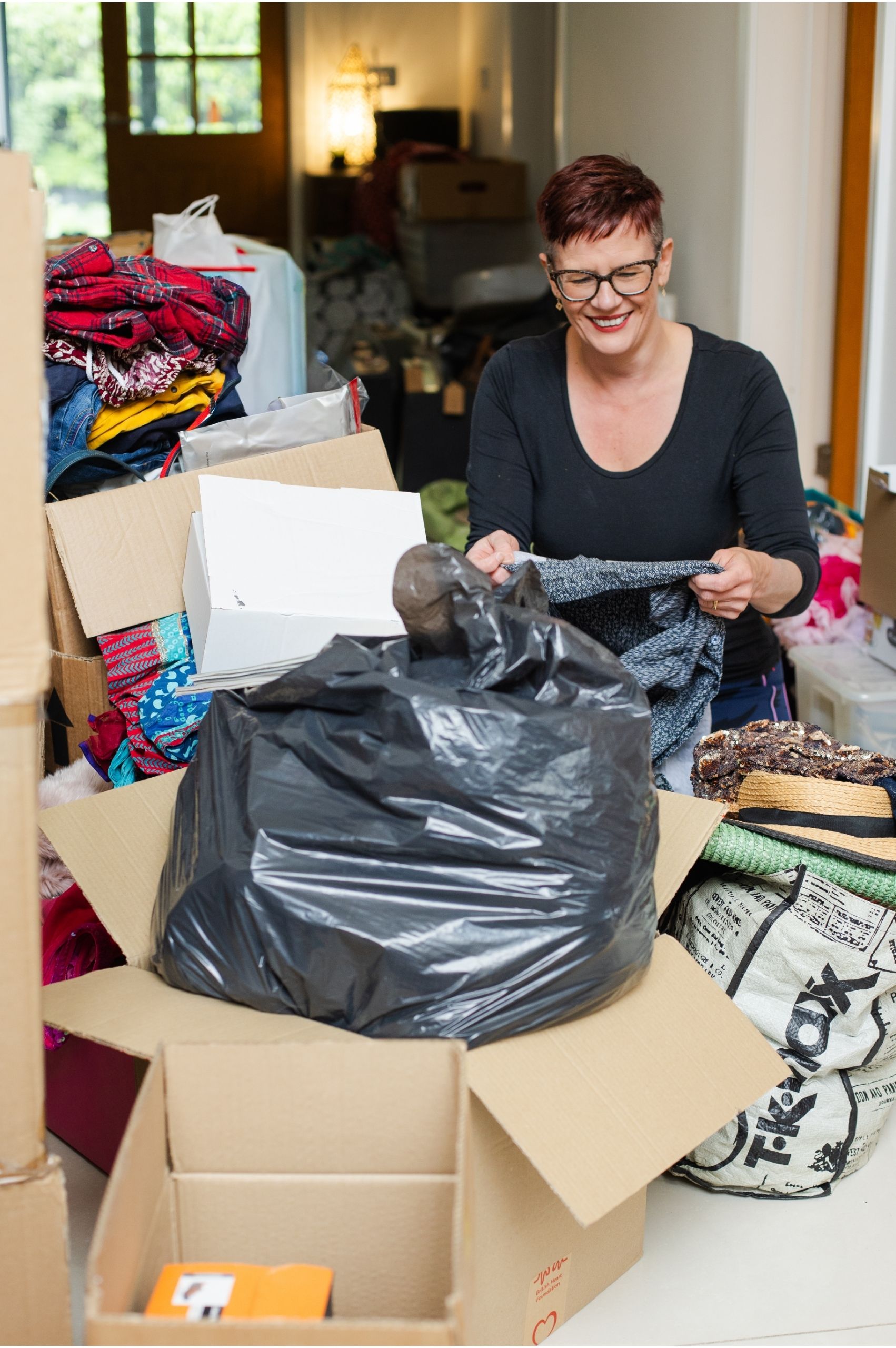 Roxy, founder of Declutter and reorganise with Roxify, sits indoors surrounded by cardboard boxes, a large black bin bag, and piles of clothing. She is wearing a black top, smiling warmly, and holding up a piece of grey clothing as she works through the sorting process.
